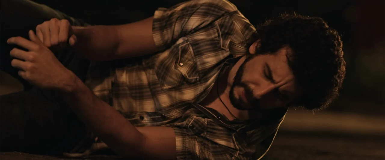 Kimura (2017) movie still: medium — A dark outdoor space with a paved surface dimly lit by a nearby light. A man lies on the ground with his eyes…