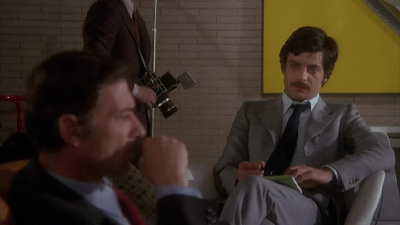 The Black Belly of the Tarantula (1971) movie still: medium — An interior space with a brick wall and a prominent yellow rectangular art piece. One…