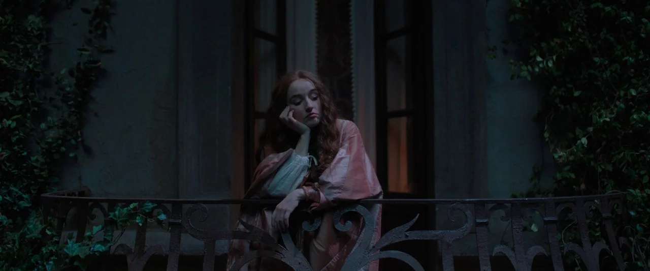 Rosaline (2022) movie still: medium — A stone balcony at night features iron railings draped with creeping ivy plants. A young woman with long…