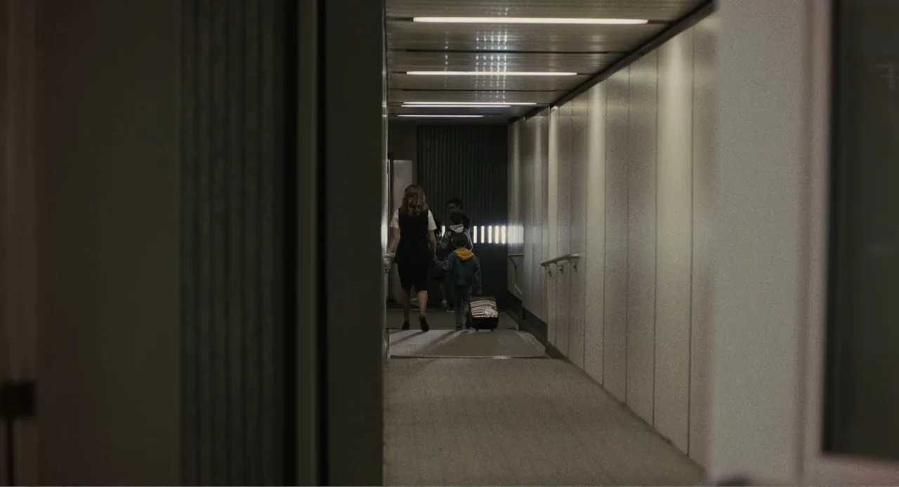 Beautiful Boy (2011) movie still: fullbody — A long, windowless airport jet bridge with white panelled walls and carpeted flooring. A woman and two…