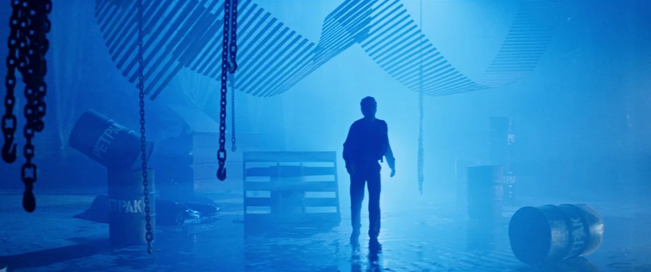 Razorback (1984) movie still: wide — An industrial storage room filled with blue haze, hanging chains, and metal barrels. A man walks through the…
