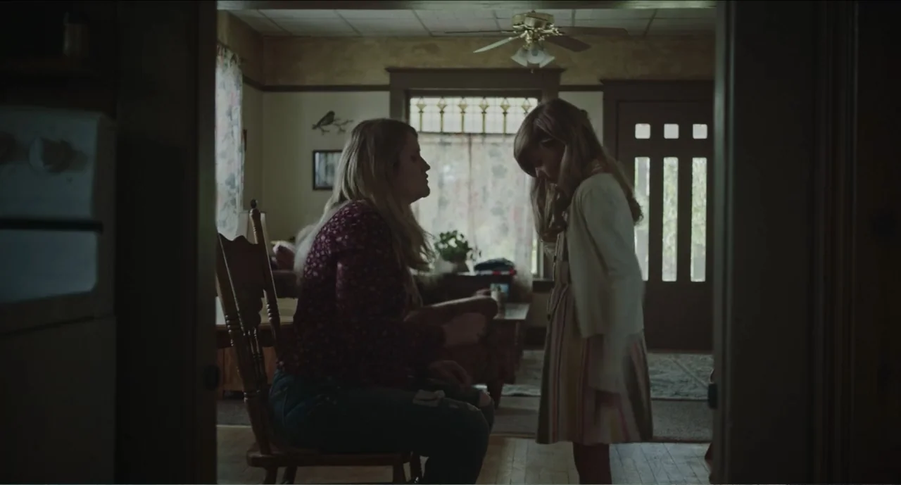 Cowboys (2014) movie still: medium — A rustic interior features a wood-framed window, wooden chairs, and simple wall decor. A seated woman faces a…