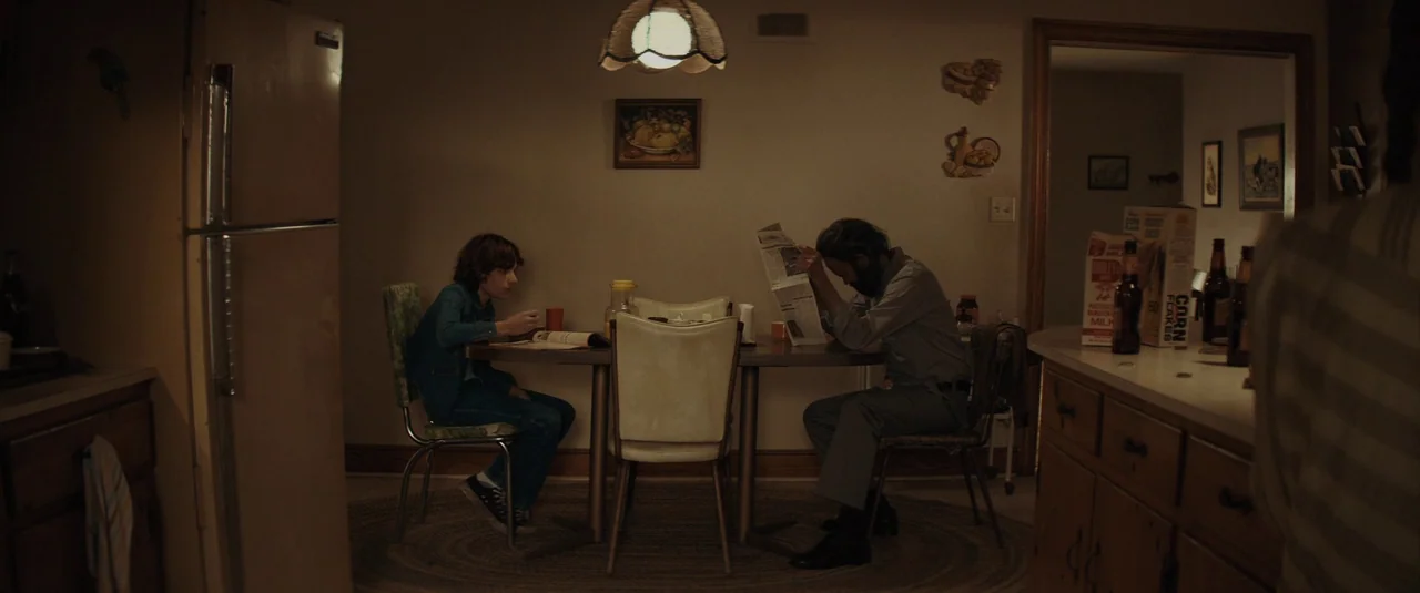 The Black Phone (2021) movie still: wide — A 1970s interior with brown wood cabinets and yellow-toned walls. Two people sit across from each other…