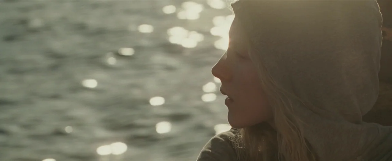 Hanna (2011) movie still: closeup — A person in a hood sits by a body of water during golden hour. The water surface reflects sunlight with high…