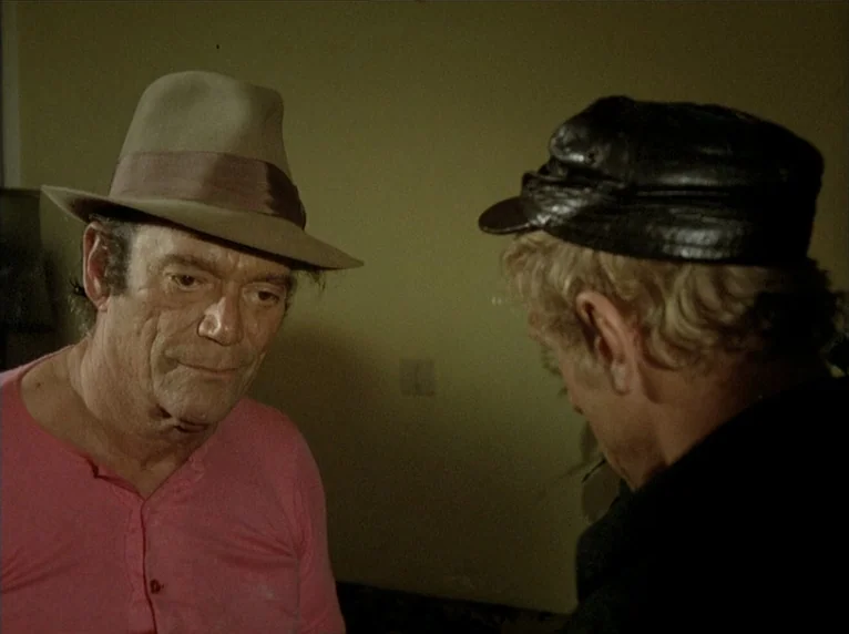 Beware Of A Holy Whore (1971) movie still: medium — Two men stand close together in a room with plain, mustard-colored walls. One man wears a tan…