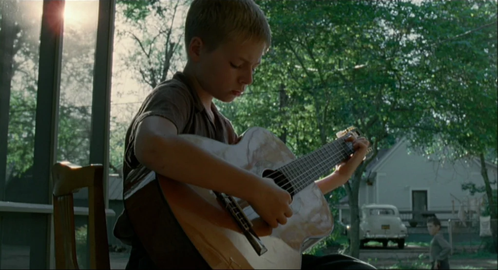 The Tree Of Life (2011) movie still: medium — A rural porch overlooking a yard with a vintage car and trees. A young boy plays an acoustic guitar…