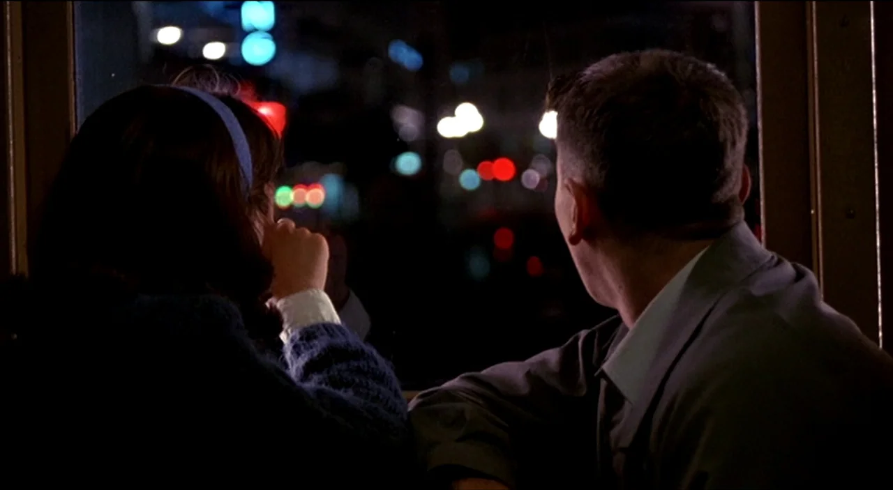 Dogfight (1991) movie still: medium — A man and a woman look out a window from inside a dark vehicle. The city lights appear as out-of-focus bokeh…