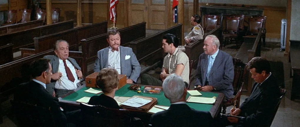 Wild in the Country (1961) movie still: wide — A wooden paneled room with rows of empty benches and American flags behind. Several people sit around…