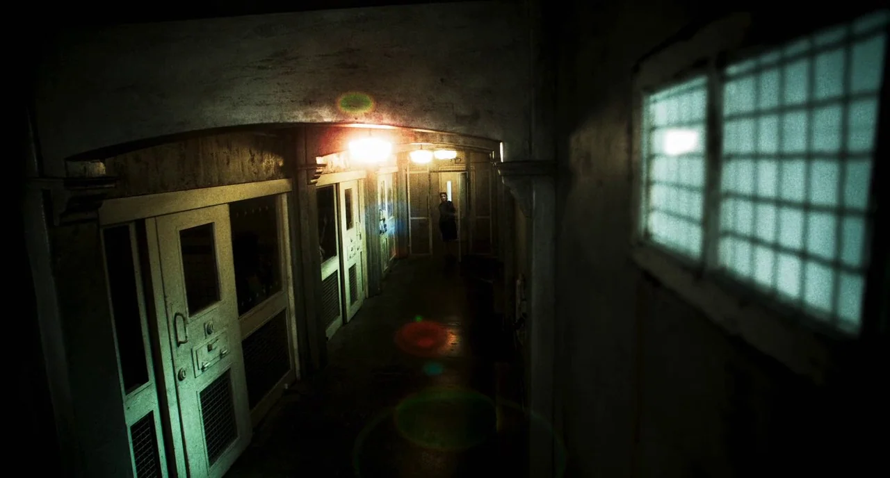 3 From Hell (2019) movie still: wide — A long, dimly lit prison hallway features heavy metal cell doors and barred windows. The narrow passage is…