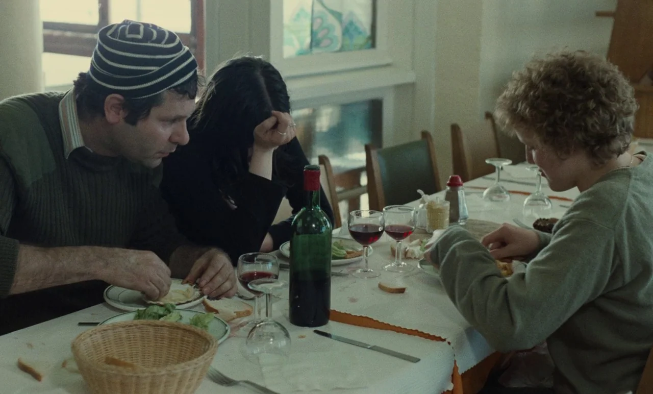 Graduate First (1978) movie still: medium — A dining table is covered with a white cloth, plates, glasses, and a wine bottle. Three people sit…