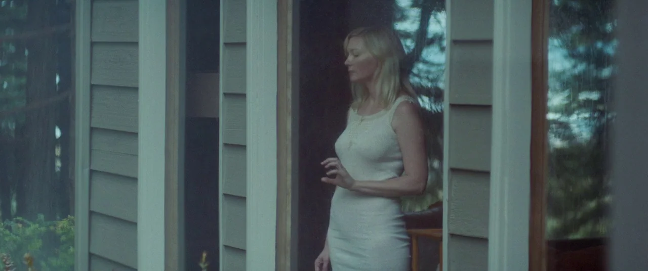 Woodshock (2017) movie still: medium — A woman stands in the doorway of a house with light grey wooden siding. She gazes outward while holding her…