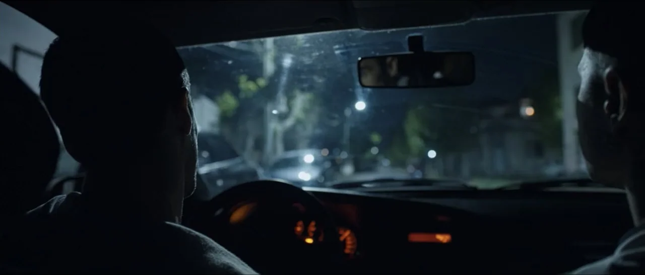 Gunman (2025) movie still: medium — Inside a dark vehicle at night with blurred street lights visible through the windshield. Two men sit in the…