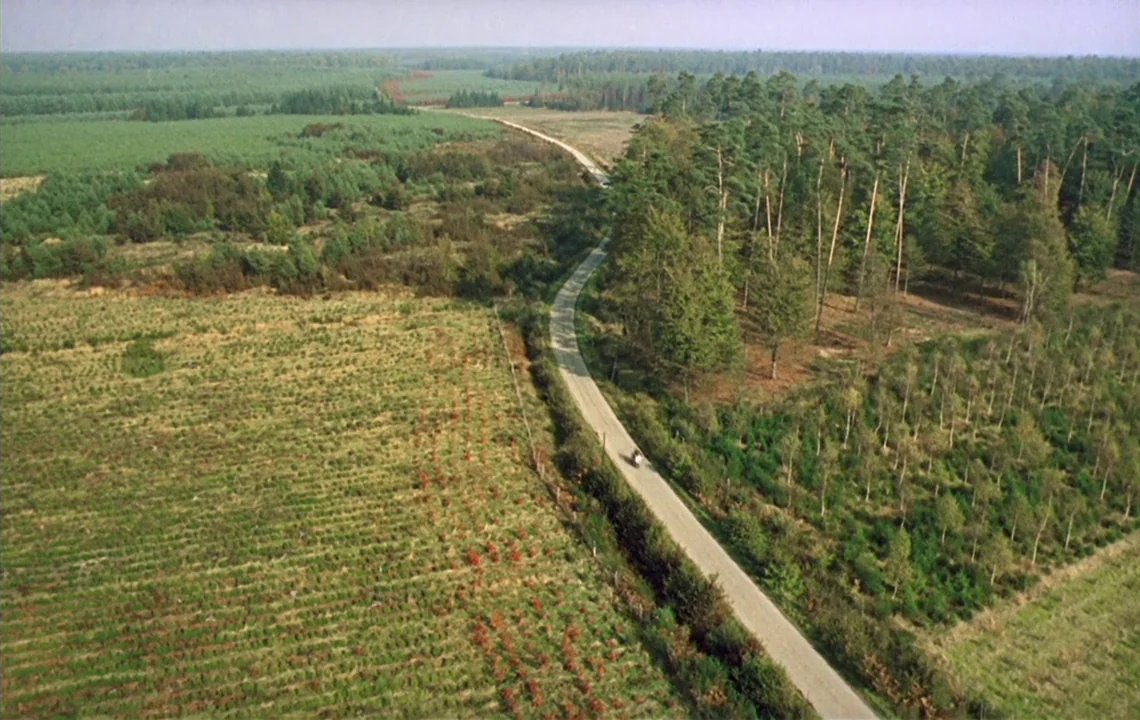 The Girl on a Motorcycle (1968) movie still: establishing — A winding paved road cuts through a landscape of dense pine trees and farmland. A lone…