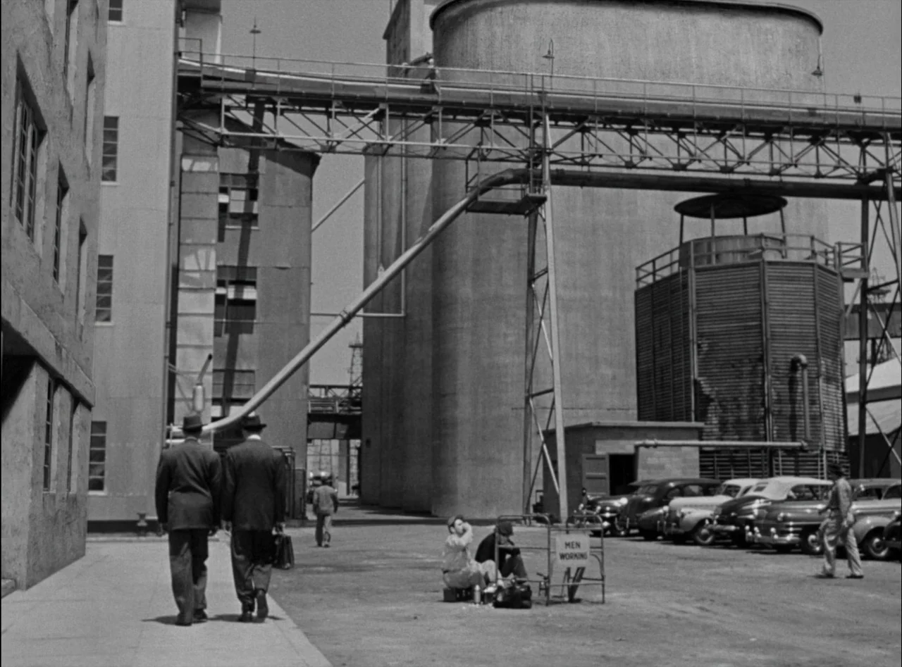 Criss Cross (1949) movie still: wide — An industrial facility features large concrete silos and metal walkways under a clear sky. Two men in suits…