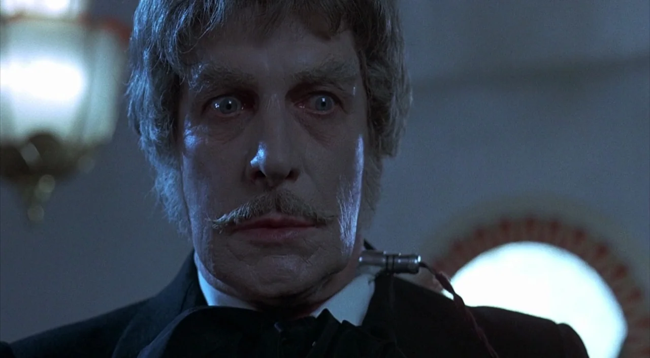 Dr. Phibes Rises Again (1972) movie still: closeup — A dimly lit interior space features an older man with a thin mustache. He stares ahead with…