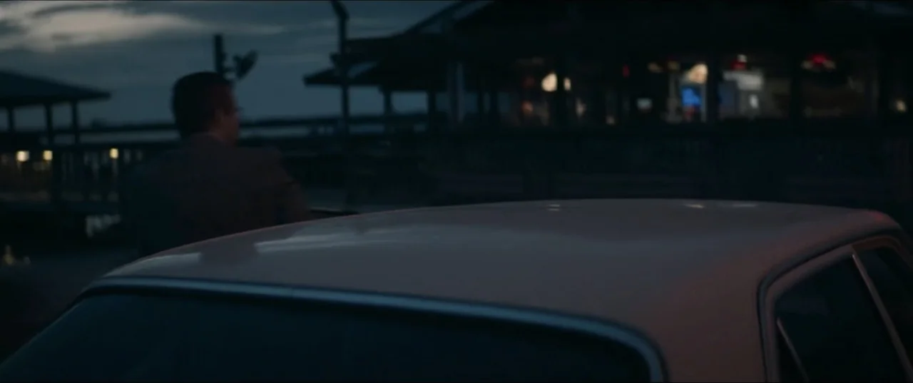 Offseason (2022) movie still: medium — A dark outdoor setting at dusk featuring a parked car in the foreground. A man stands near the vehicle while…