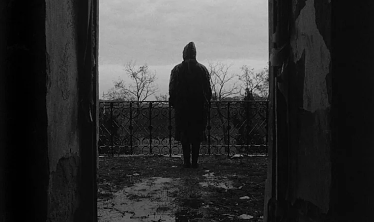 Sátántangó (1994) movie still: fullbody — A figure stands on a balcony at the edge of a crumbling interior space. The person looks out toward bare…