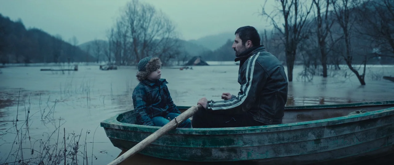R.M.N. (2022) movie still: wide — A man and a boy sit in a weathered rowboat atop a vast flooded landscape. Bare, leafless trees rise from the murky…