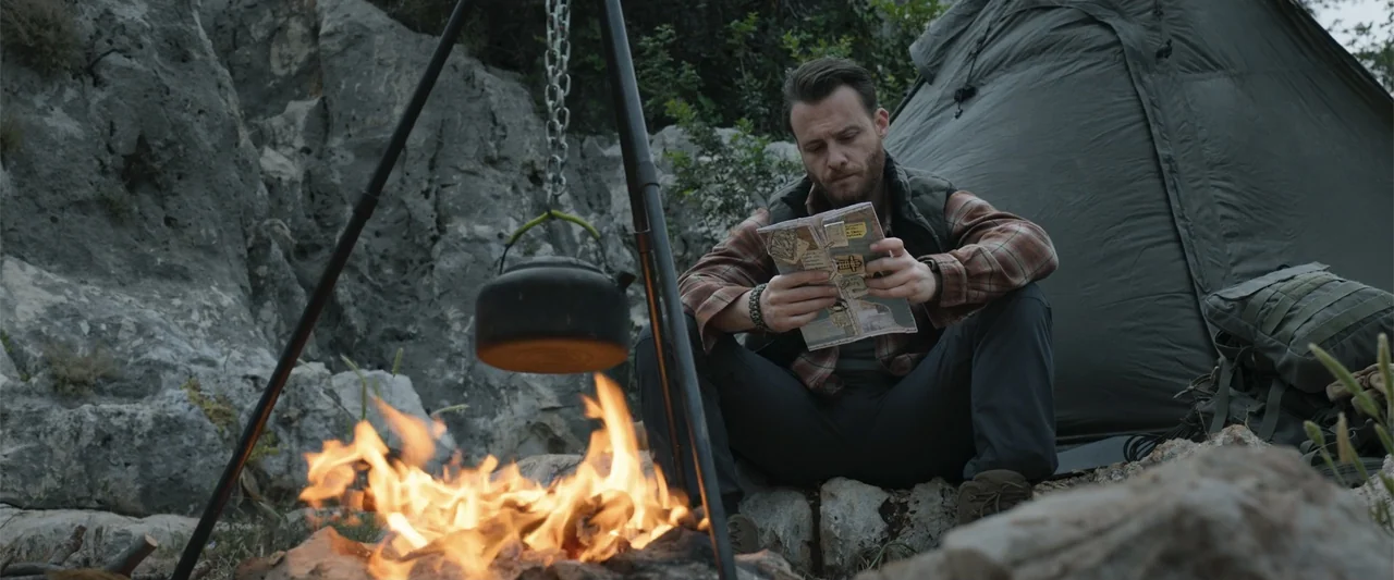 Blue Cave (2024) movie still: medium — A rocky campsite at night featuring a tent and a tripod over a fire. A bearded man sits by the fire while…