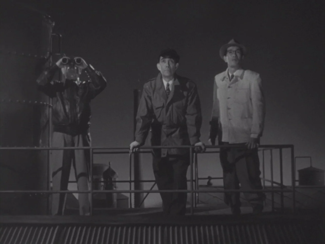 Godzilla Raids Again (1955) movie still: fullbody — Three men stand on a rooftop at night behind a metal railing. They observe the sky while leaning…