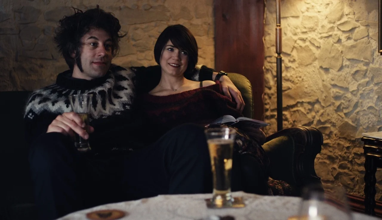 Land Ho! (2014) movie still: medium — A living room with light-colored stone walls and a dark sofa. A man and a woman sit together holding glasses…
