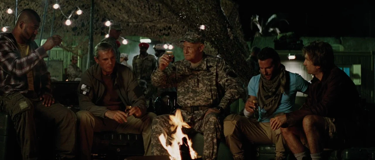 The A-Team (2010) movie still: wide — A group of men in tactical and casual clothing sit around a low campfire at night. The campsite is surrounded…