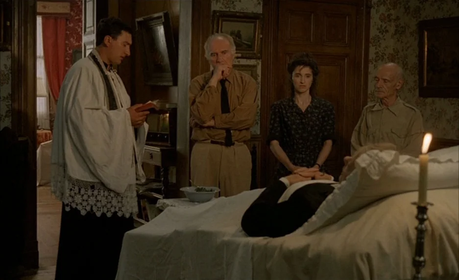 Milou en Mai (1990) movie still: medium — A traditional room with floral wallpaper and framed artwork houses a deathbed scene. A priest reads from a…