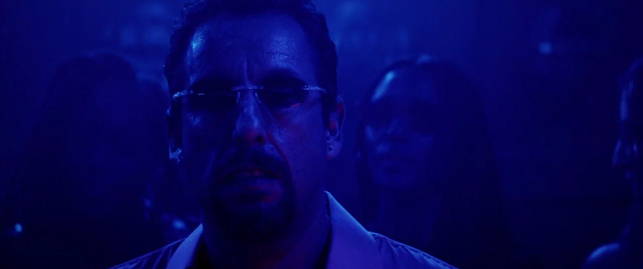 Uncut Gems (2019) movie still: closeup — A man stands in a dark environment illuminated only by harsh blue artificial light. Other figures are…