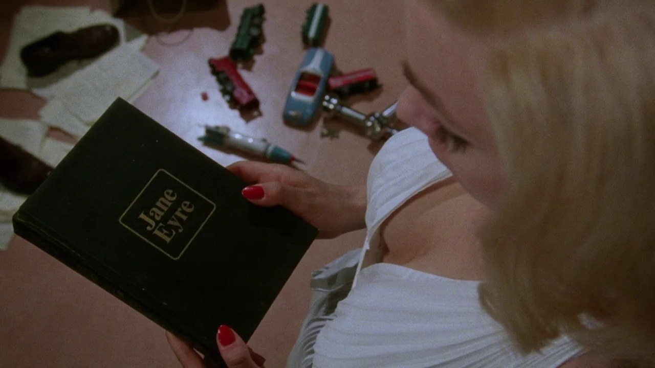 Insignificance (1985) movie still: medium — A woman stands in a room with scattered children's toy cars and papers. She holds a copy of the book…