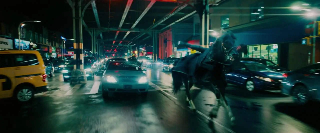 John Wick: Chapter 3 – Parabellum (2019) movie still: wide — An urban multi-lane road runs underneath a dark steel elevated train structure at…