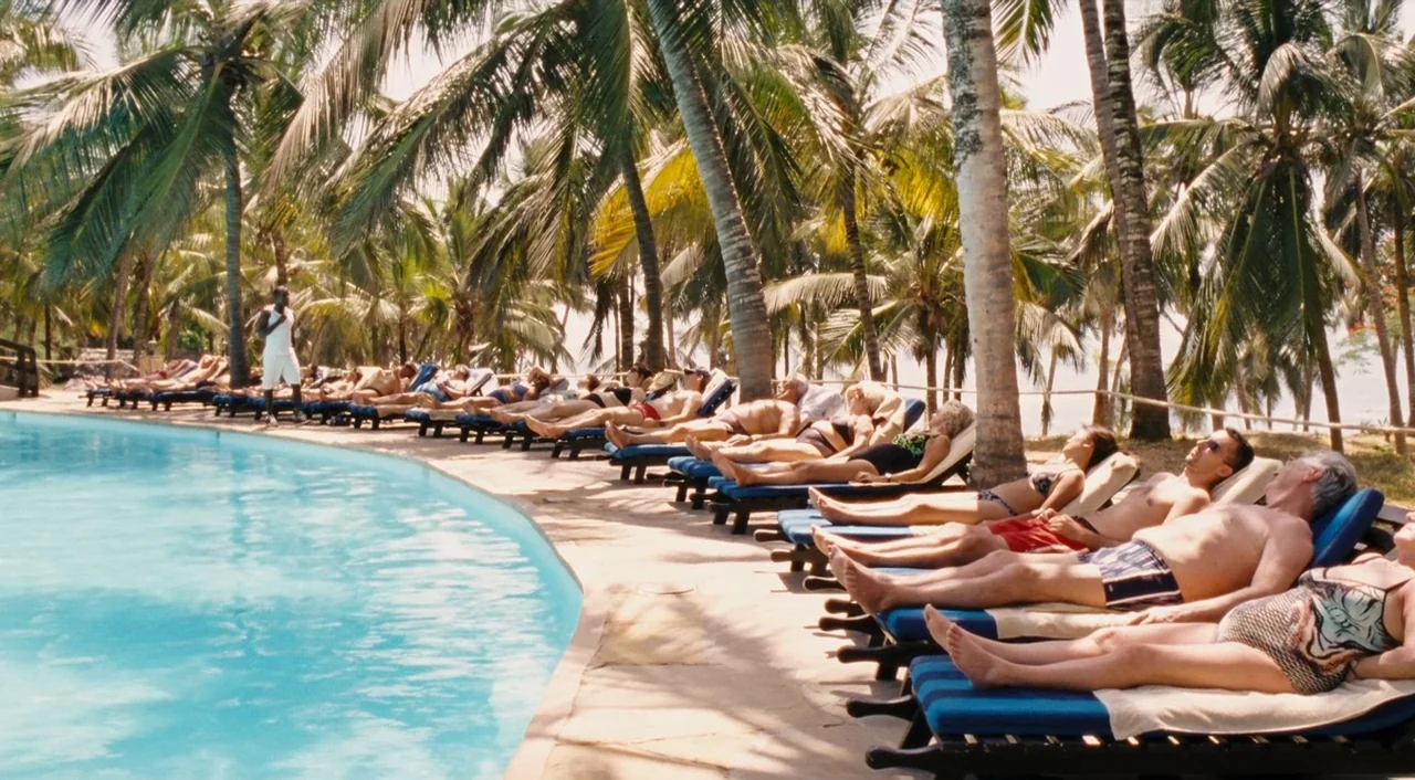 Paradise: Love (2012) movie still: wide — A curved swimming pool edge is lined with numerous people sunbathing on blue loungers. Tall palm trees…
