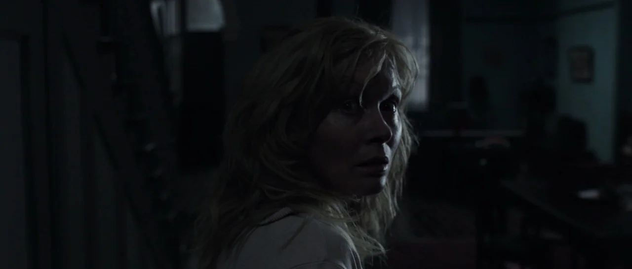 The Babadook (2014) movie still: closeup — A dark house interior with minimal visible details in the background shadows. A woman looks toward the…