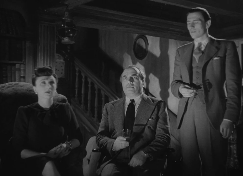 Dead of Night (1945) movie still: medium — A dimly lit room features a staircase, bookshelves, and a hanging lantern lamp. Three people are seated…