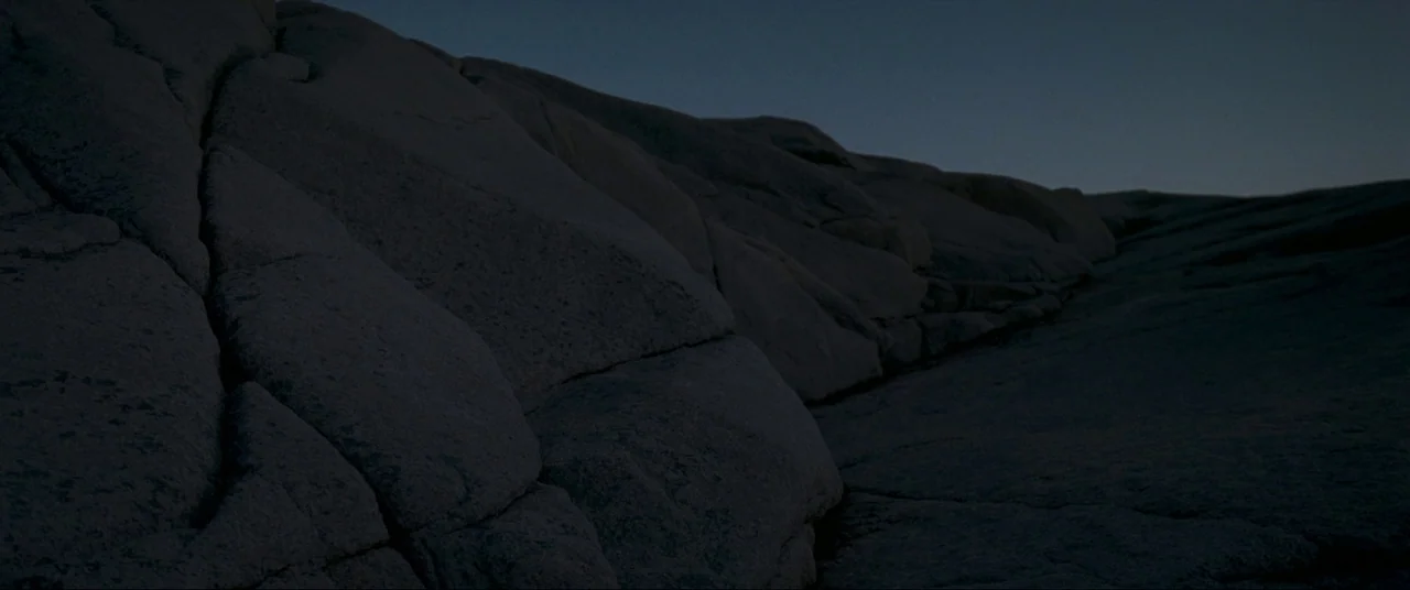 Leviathan (2014) movie still: establishing — A dark, textured stone formation stretches across the frame against a twilight sky. There are no people…