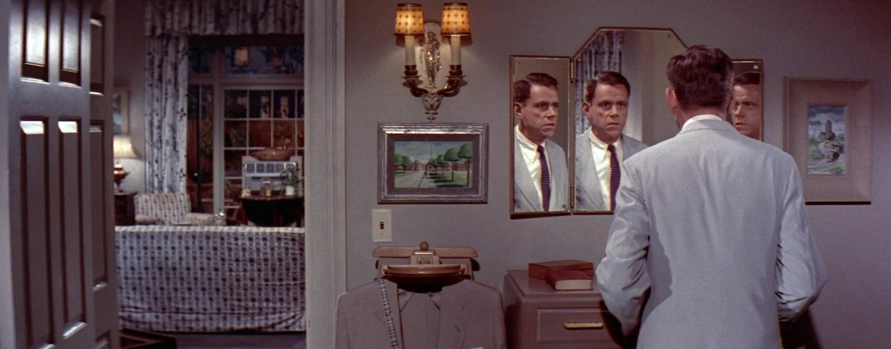 The Seven Year Itch (1955) movie still: medium — A man in a light grey suit faces a triple-panel wall mirror. Beyond the open doorway is an adjacent…