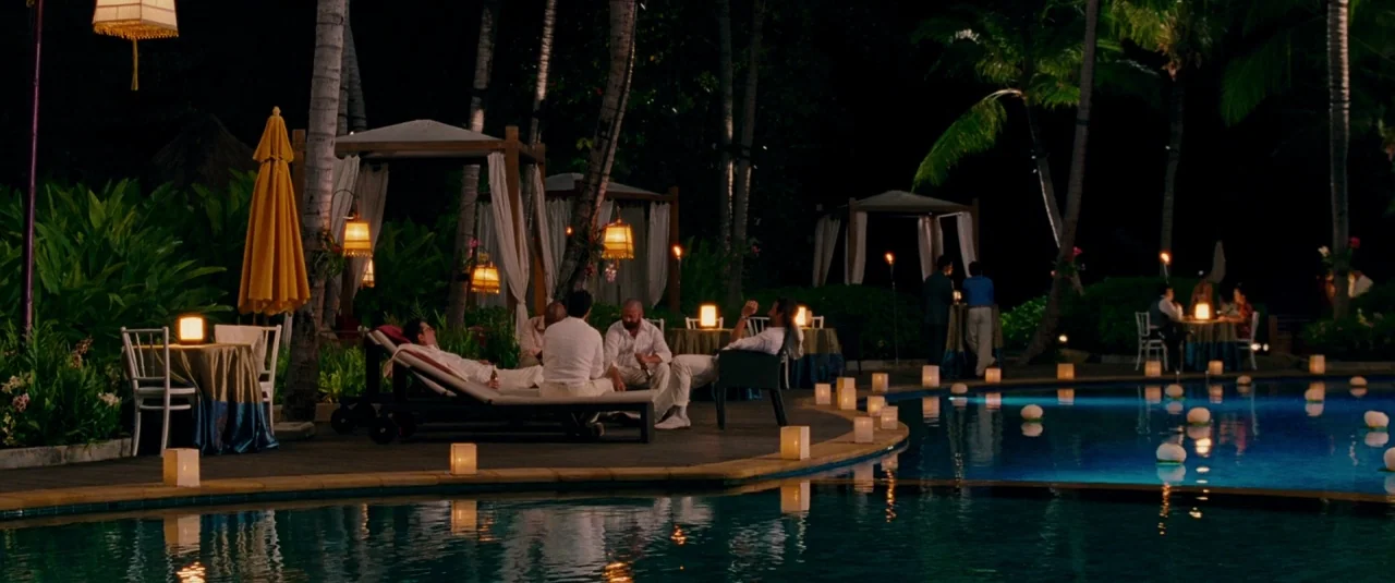 The Hangover Part II (2011) movie still: wide — A night view of a tropical hotel pool area with palm trees and cabanas. Several guests sit in white…