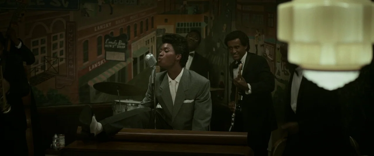 Elvis (2022) movie still: medium — A dimly lit club interior features a painted mural of storefronts on the back wall. A singer in a grey suit…