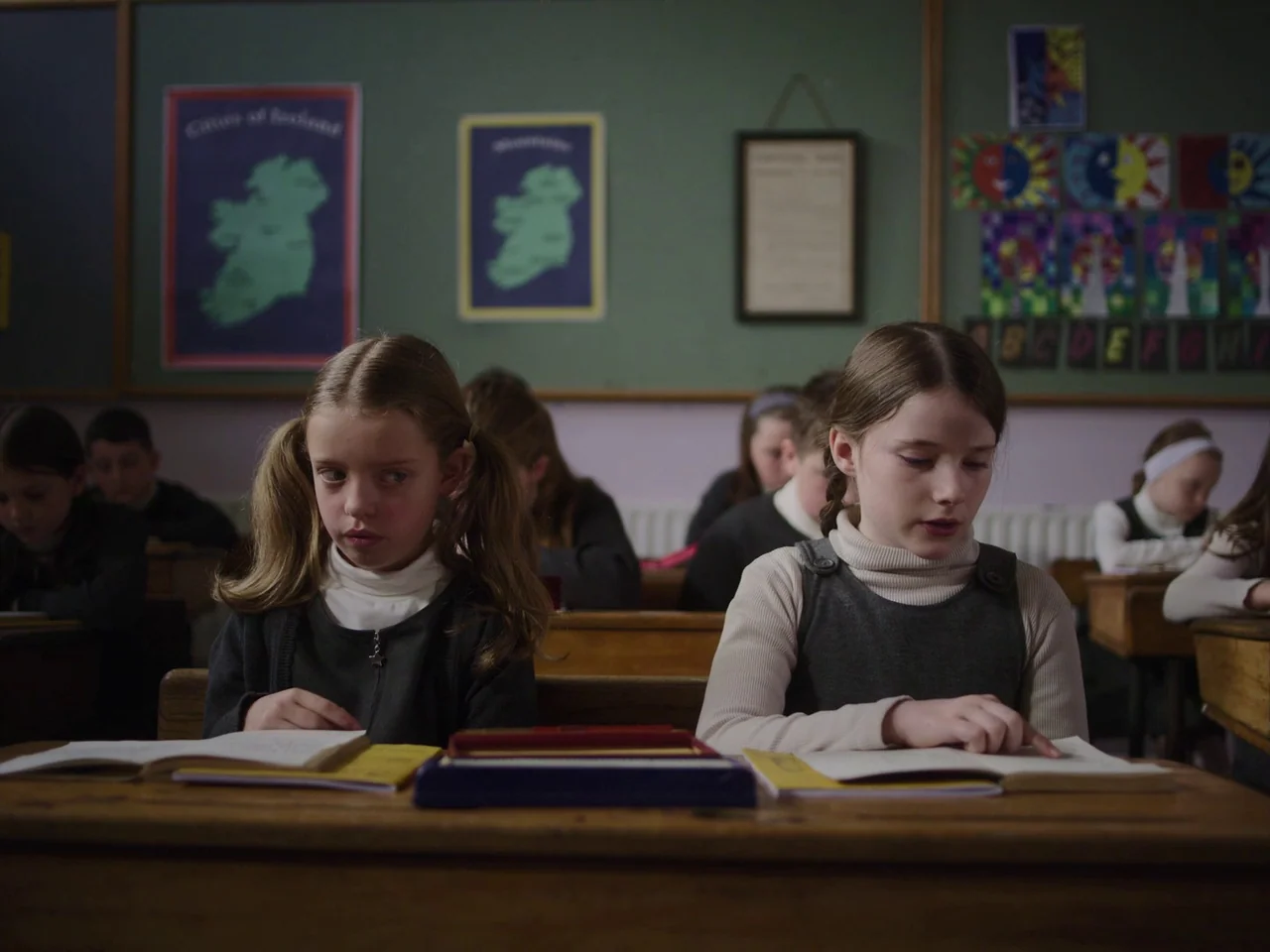 The Quiet Girl (2022) movie still: medium — A classroom features dark green walls decorated with educational posters and children's artwork. Two…