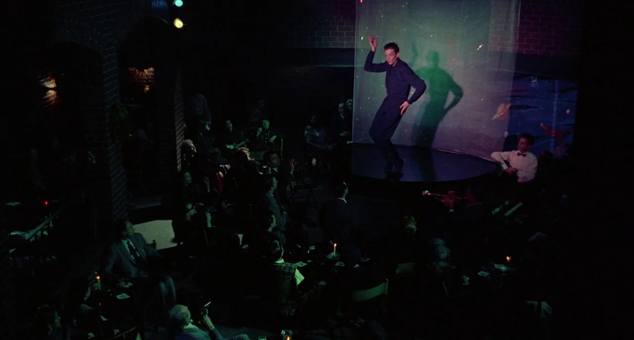 Bell Book and Candle (1958) movie still: wide — A dimly lit nightclub with brick walls and guests seated at tables in shadow. A man performs a solo…