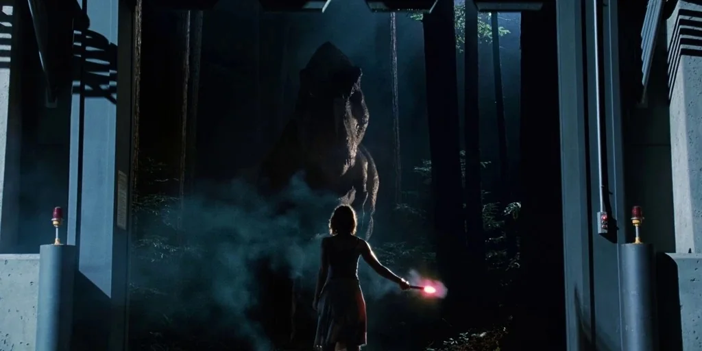 Jurassic Park (1993) movie still: wide — A woman holding a flare stands at an industrial gate threshold in a dark forest. A large dinosaur stands in…