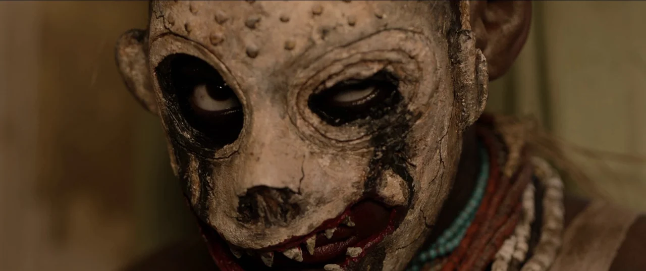 His House (2020) movie still: closeup — A close-up shot focuses on a person wearing a weathered, hand-crafted tribal mask. The figure is adorned…