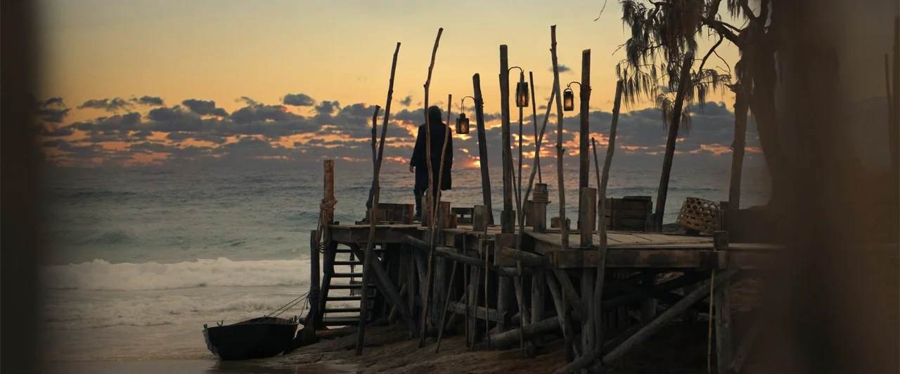 The Bluff (2026) movie still: wide — A rustic wooden pier extends into the ocean during a vibrant golden hour sunset. A lone figure stands at the…