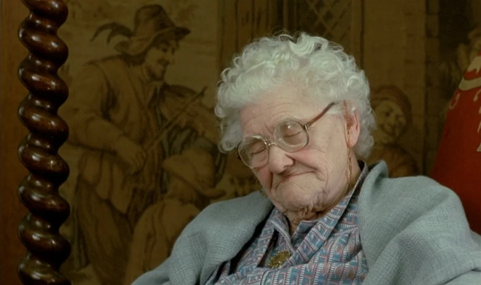 Vagabond (1985) movie still: closeup — An elderly woman sits in a chair in front of a patterned wall hanging. The subject rests with her eyes…