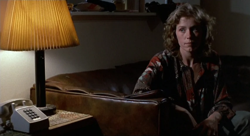 Blood Simple (1984) movie still: medium — A dimly lit room contains a leather sofa and a side table with a lamp. A woman sits on the couch facing…