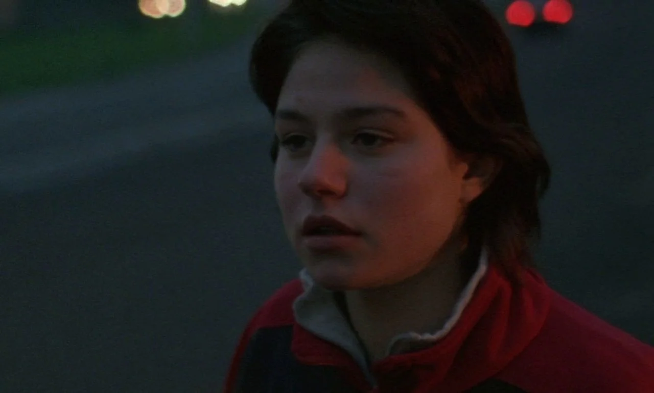 Rosetta (1999) movie still: closeup — A dark outdoor area at night with blurred distant light spots on the road. A teenage girl with short brown…