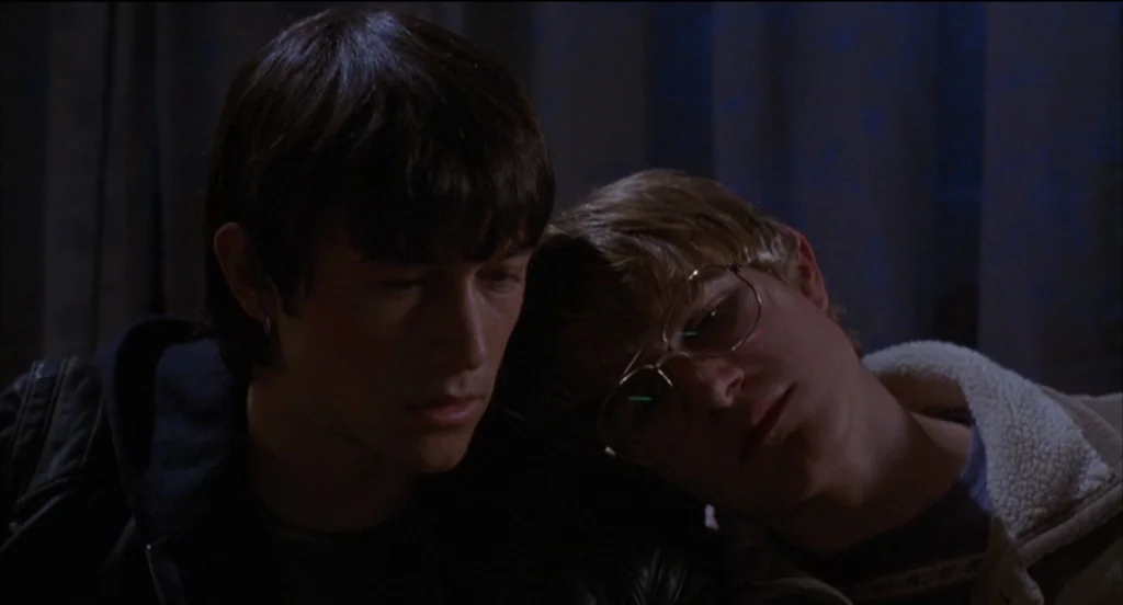 Mysterious Skin (2004) movie still: closeup — A dark indoor space with heavy curtains visible in the background. Two young men sit together, one…