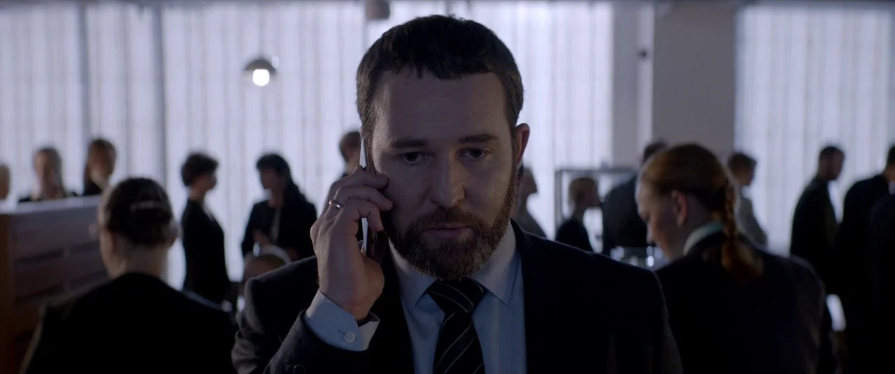 Loveless (2017) movie still: closeup — An open office space features people working at desks in the blurred background. A man in a business suit…