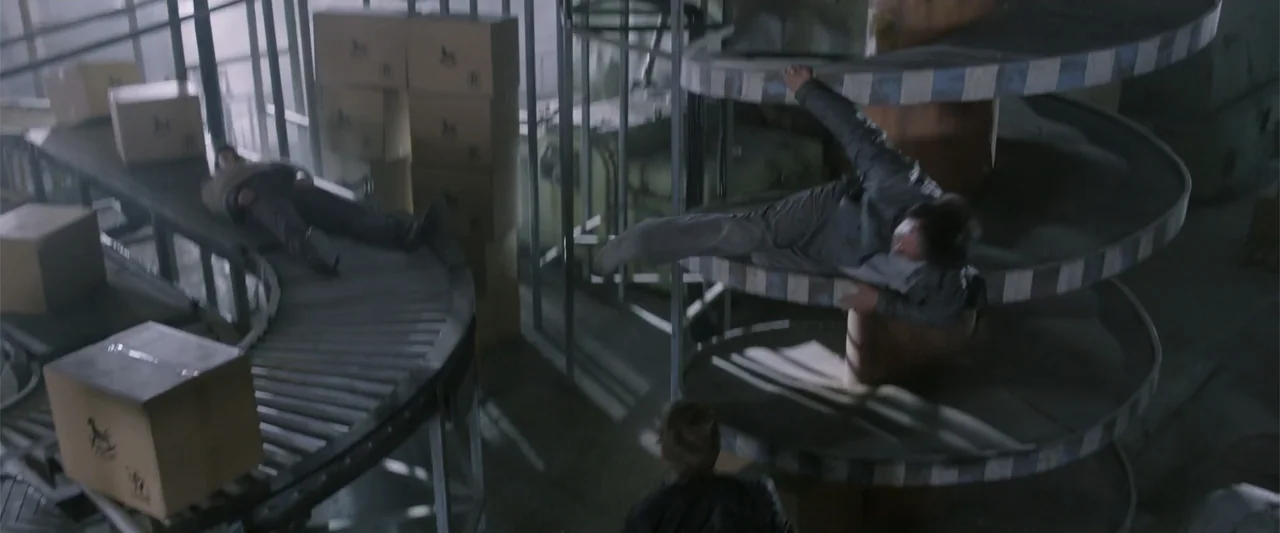 Skiptrace (2016) movie still: wide — An industrial facility features a spiral conveyor system loaded with various cardboard boxes. Three men…