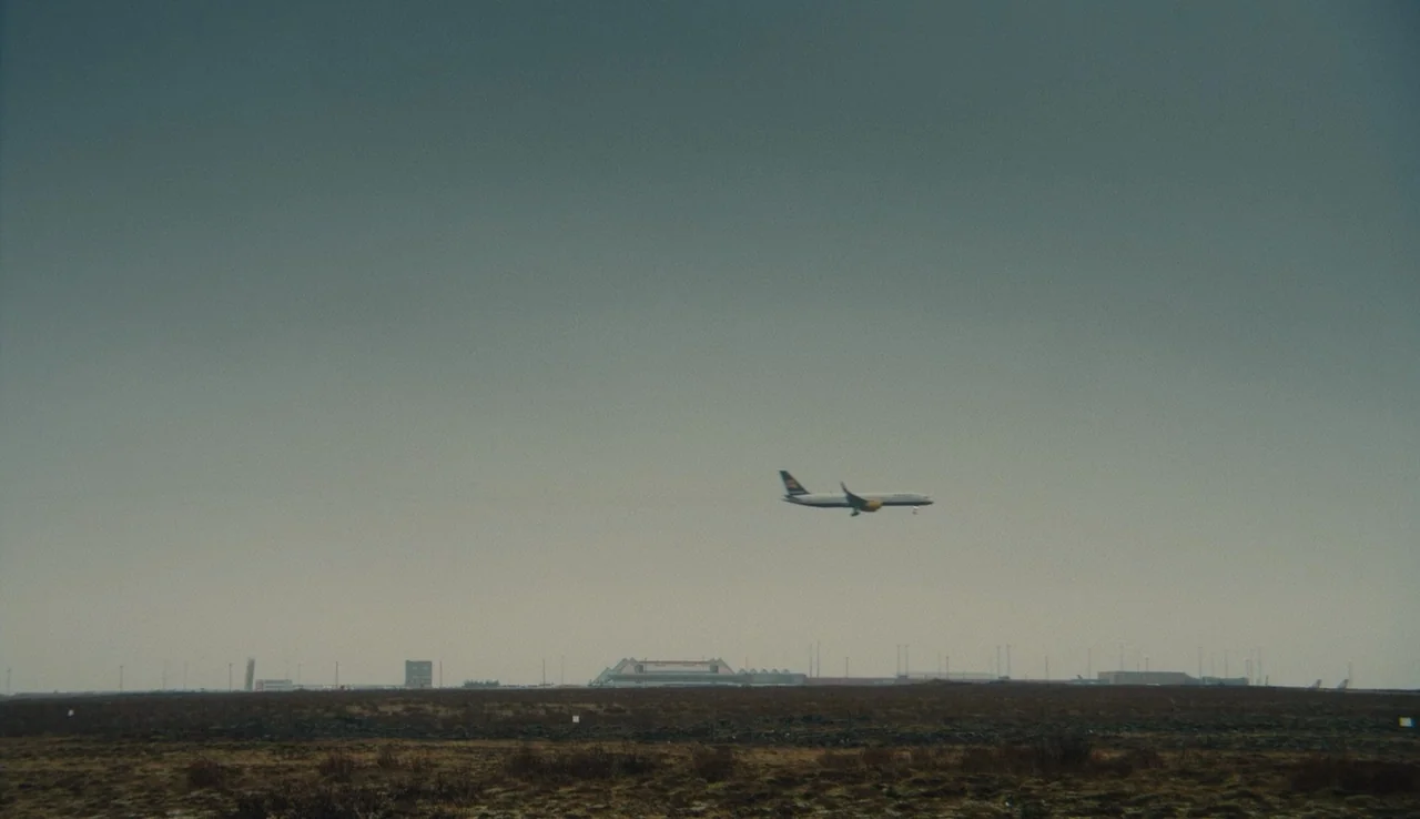 Land Ho! (2014) movie still: wide — A vast, barren landscape stretches toward a distant airport terminal under a hazy sky. A single commercial…