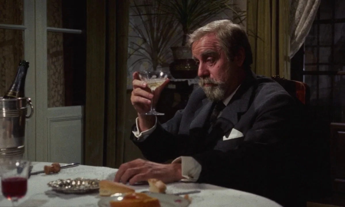 Tristana (1970) movie still: medium — A dimly lit dining room features a table set for dinner with wine. An elderly man sits at the table holding a…
